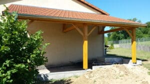 Carport en bois avec toit en tuiles rouges adossé à une maison, remorque et brouette dans un jardin.