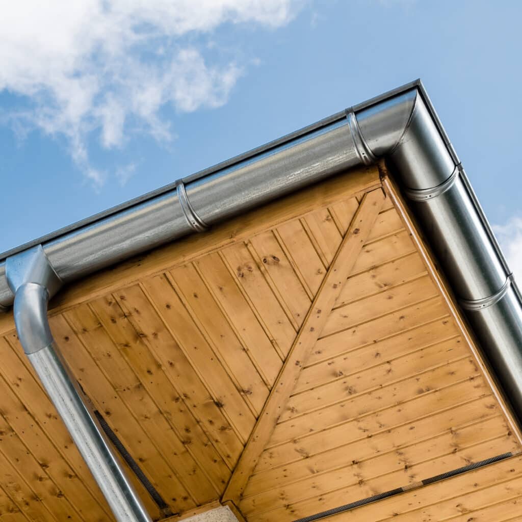 Gouttière métallique et débord de toit en bois clair Gouttière en zinc et descente pluviale sur une toiture avec sous-face en bois clair sous un ciel bleu.