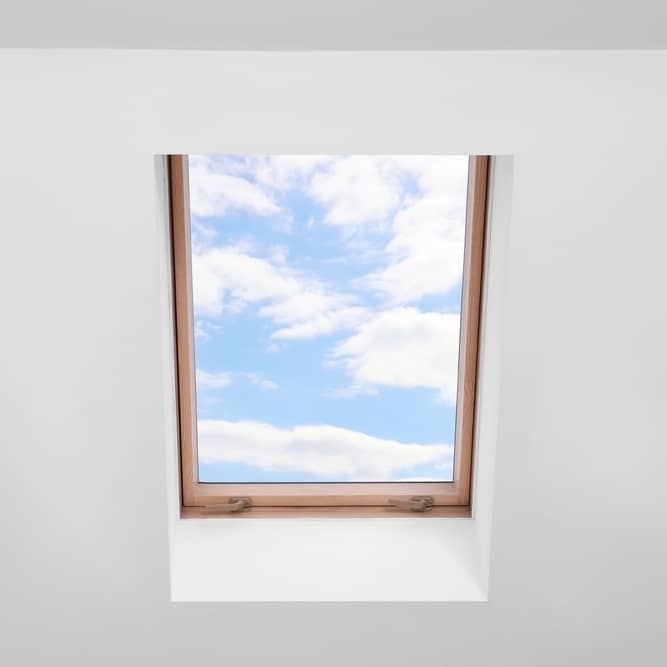 Lucarne en bois : vue panoramique sur le ciel bleu Lucarne en bois encastrée dans un plafond blanc offrant une vue dégagée sur un ciel bleu parsemé de nuages.